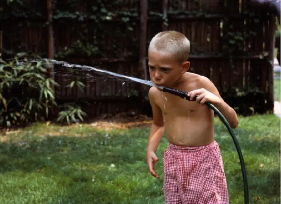 A young boy with short blond hair, wearing red checkered shorts, drinks water from a garden hose in a grassy backyard on a sunny day.