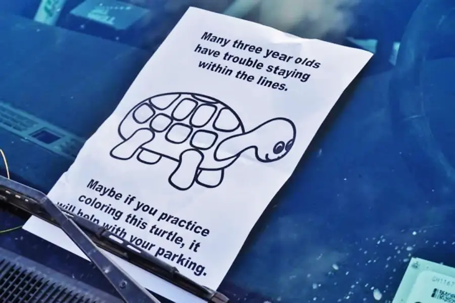 A paper with a turtle coloring page and a sarcastic note about bad parking is tucked under a car's windshield wiper. The note suggests practicing coloring to help with parking skills.
