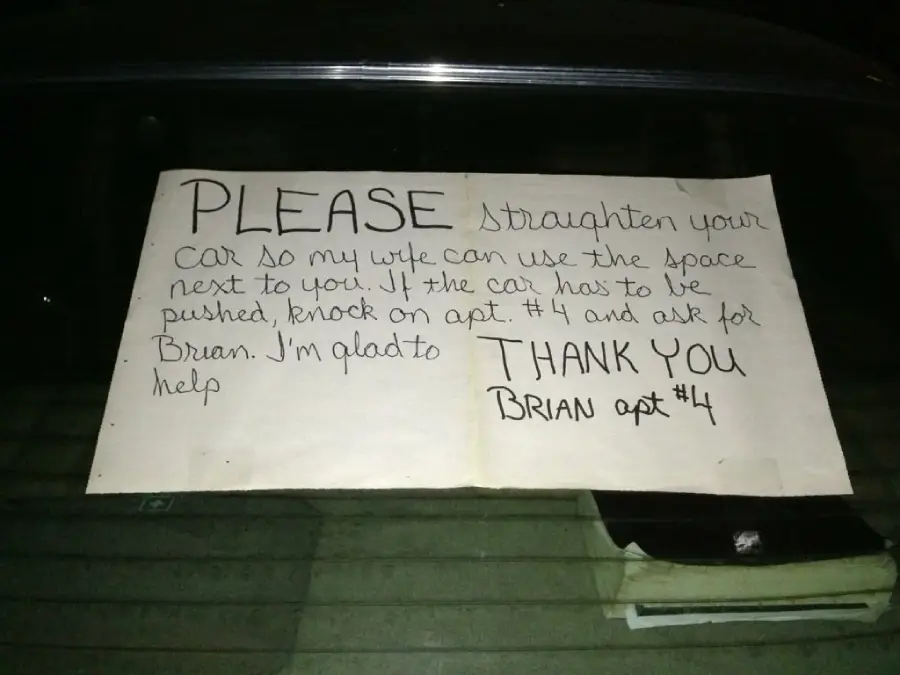 A handwritten sign on a car’s rear window asks, "Please straighten your car so my wife can use the space next to you." It gives instructions to contact Brian in apartment #4 if help is needed.