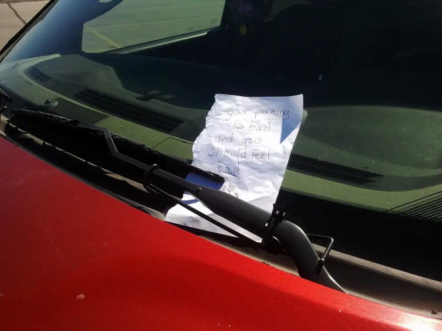A handwritten note is placed under the windshield wiper of a red car. The note reads, "Your parking is bad and you should feel bad.