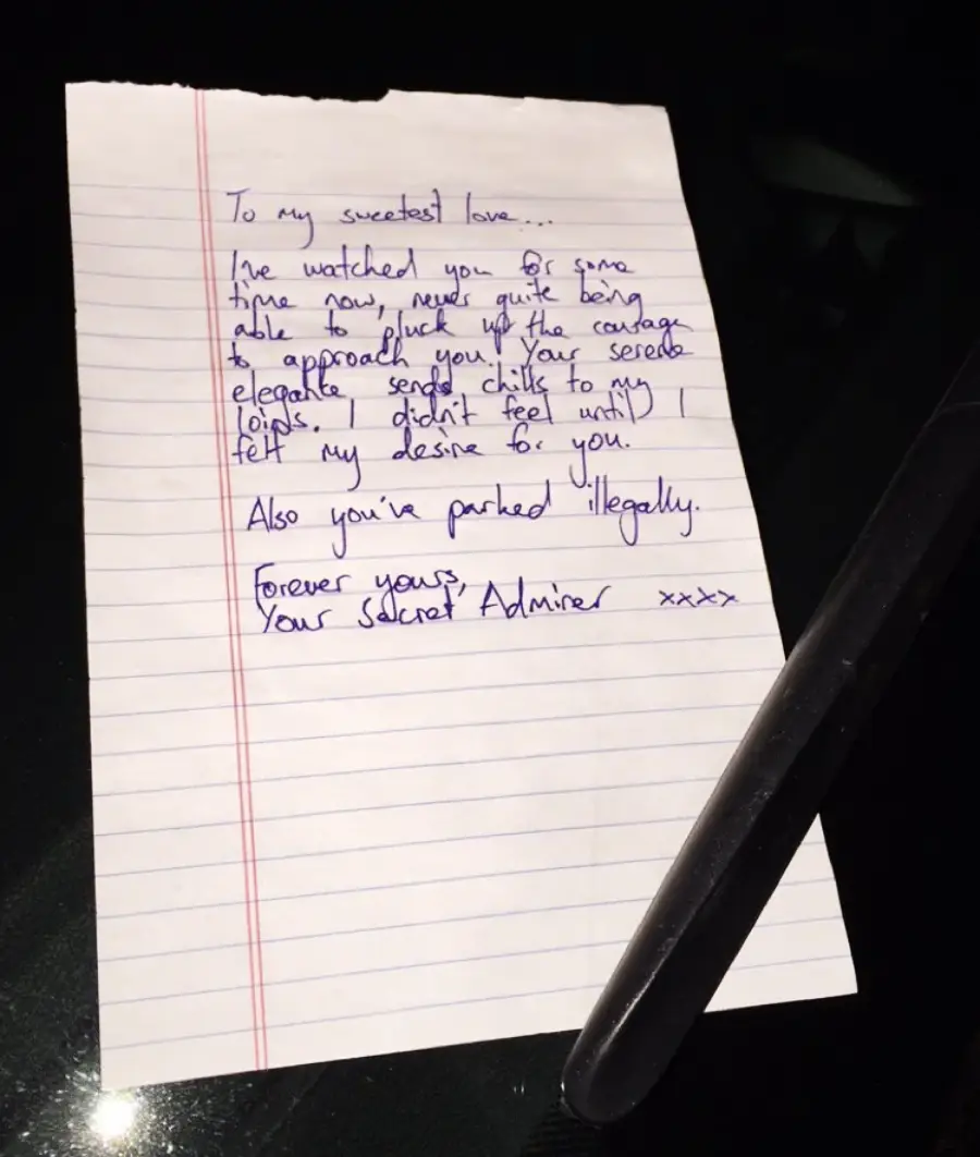 A handwritten love note on lined paper sits on a table with a pen. The note expresses secret admiration, mentions watching from afar, and humorously points out the recipient has parked illegally.