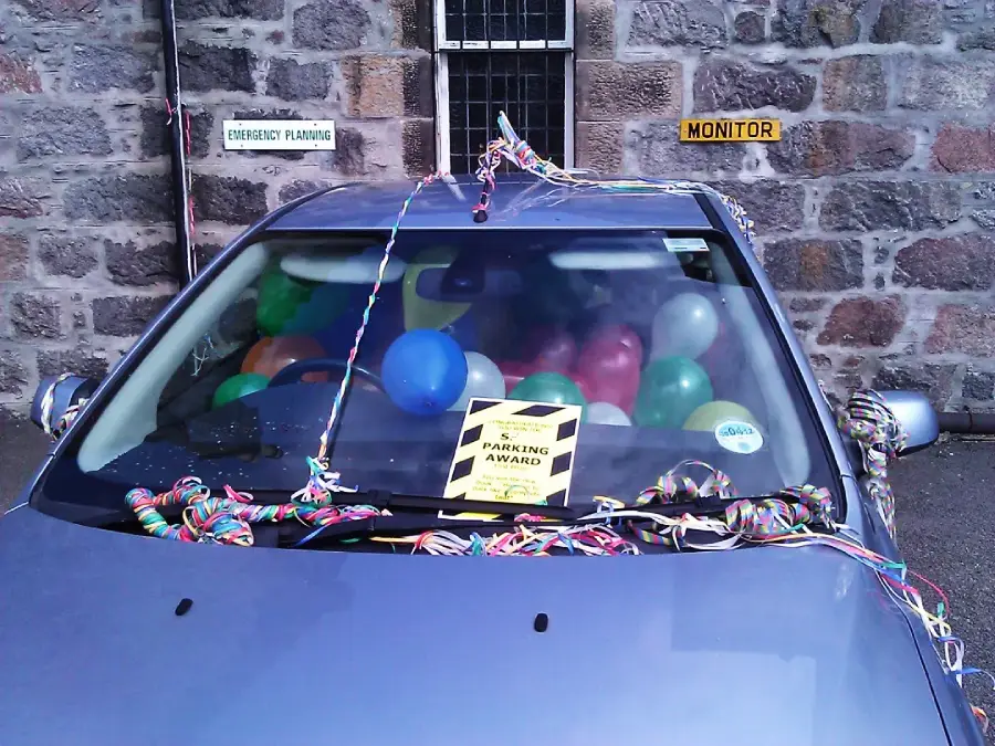 A car with its interior filled with colorful balloons and streamers is parked in front of a stone wall. A humorous "Parking Award" notice is displayed on the windshield.