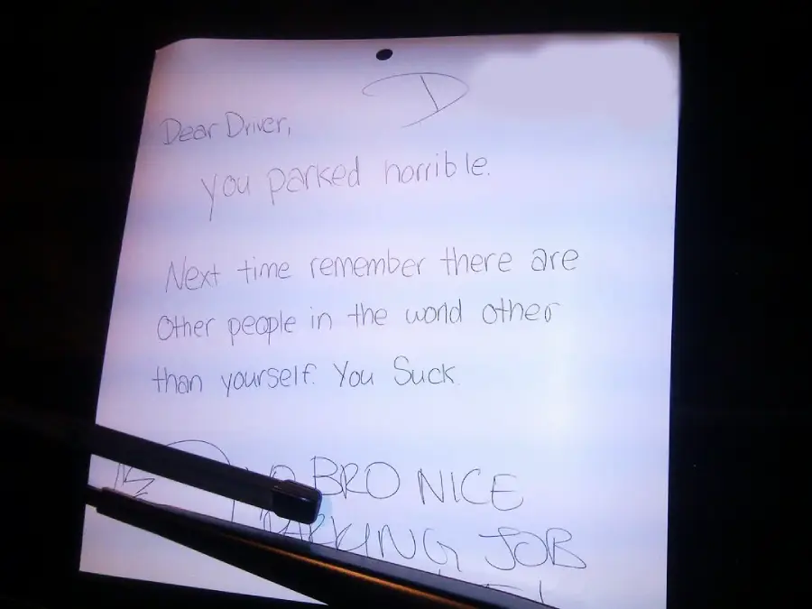 A handwritten note on white paper reads: "Dear Driver, you parked horrible. Next time remember there are other people in the world other than yourself. You suck." Pens are lying on the paper.