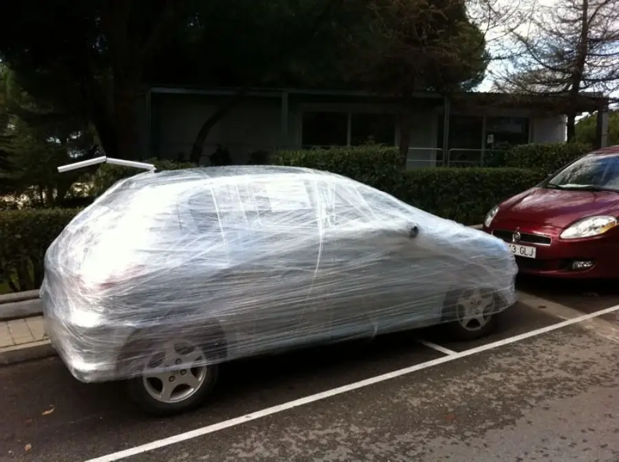 A parked car is tightly wrapped in clear plastic wrap, covering the entire vehicle. It is parked beside another car in an outdoor lot with bushes and trees in the background.