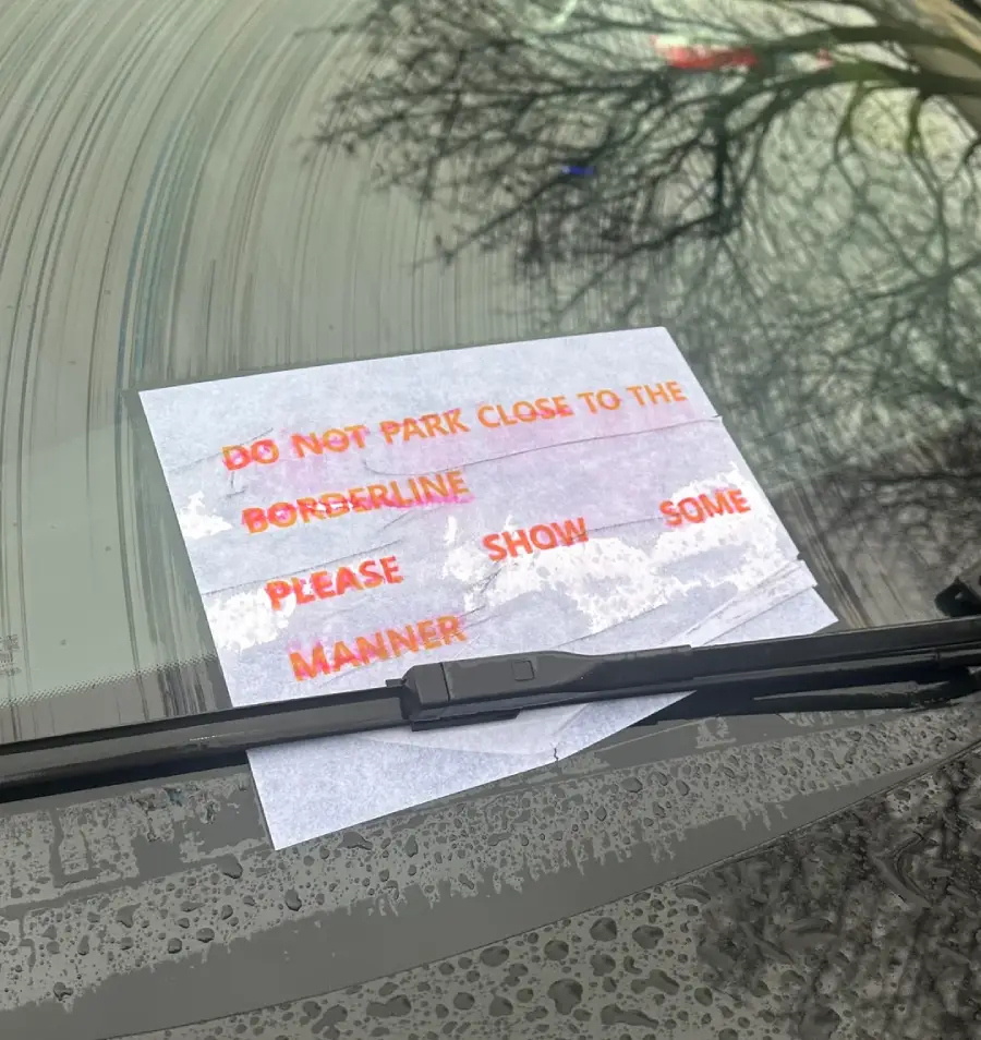 A note under a car windshield wiper reads, "DO NOT PARK CLOSE TO THE BORDERLINE. PLEASE SHOW SOME MANNER" with orange and red text on white paper. Rain and tree branches reflect on the glass.