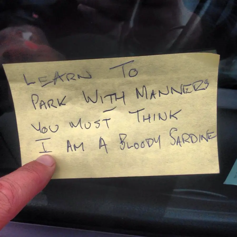 A handwritten note on a yellow sticky note reads: "Learn to park with manners. You must think I am a bloody sardine." A finger is holding the note against a car window.