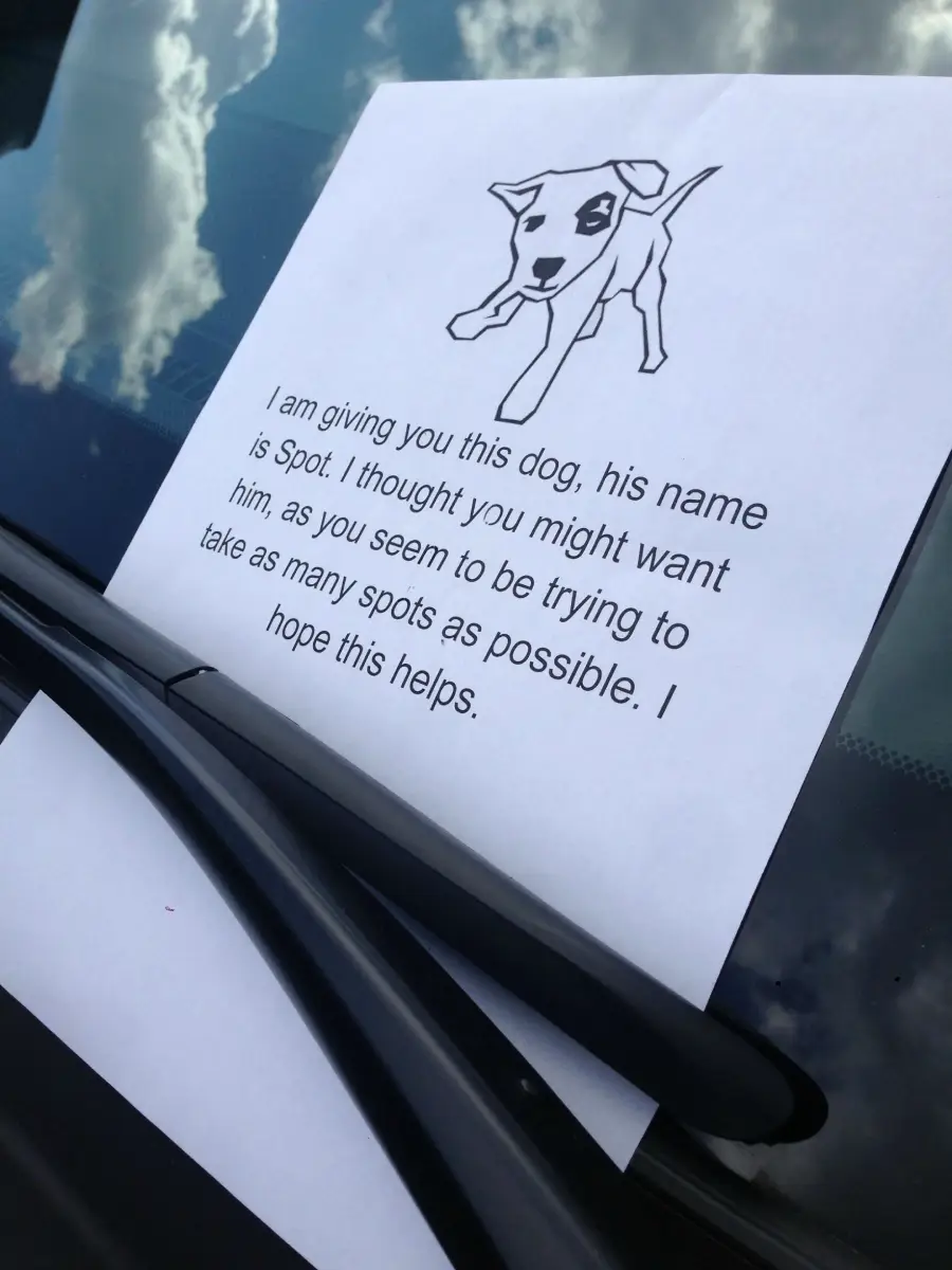 A note with a drawing of a dog named Spot is placed under a car's windshield wiper. The note sarcastically addresses the car owner for taking up multiple parking spots. Clouds are reflected on the windshield.