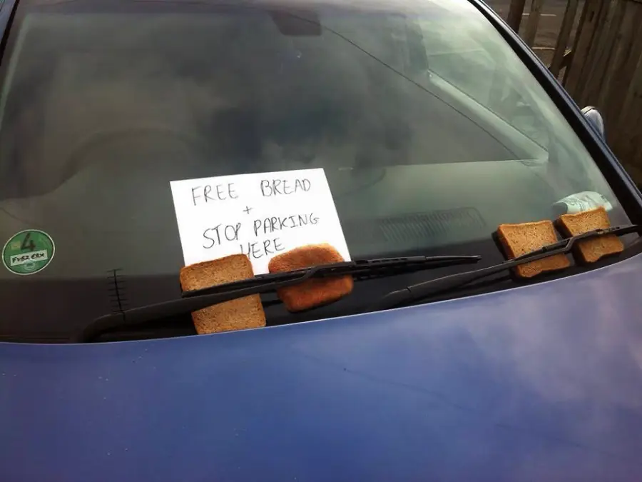 Slices of bread are placed under the windshield wipers of a blue car. A handwritten sign on the windshield reads, "FREE BREAD + STOP PARKING HERE.