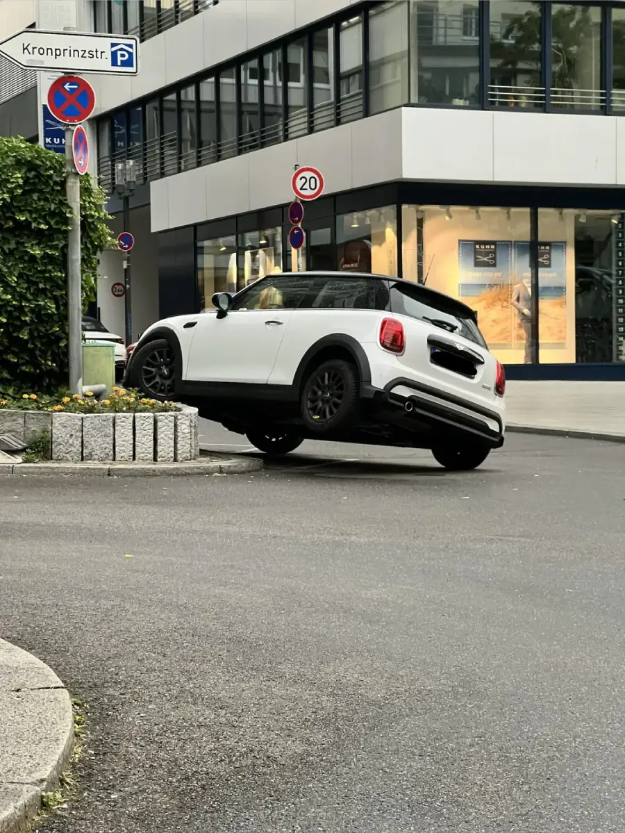 A white Mini Cooper is stuck with its rear wheels lifted off the ground on a raised curb at the corner of a city street near a building with glass windows.