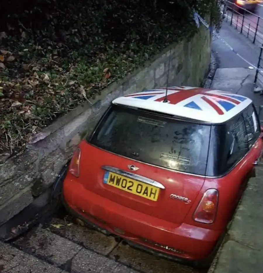 A red Mini Cooper with a Union Jack roof is wedged tightly between a stone wall and an embankment, stuck off the side of a narrow street. Its rear license plate reads "MW02 OAH.