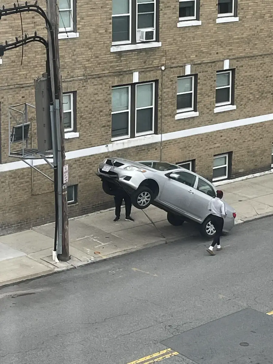 A silver car is oddly positioned with its rear end lifted against a brick building, as two men stand nearby on the sidewalk, appearing puzzled by the situation.