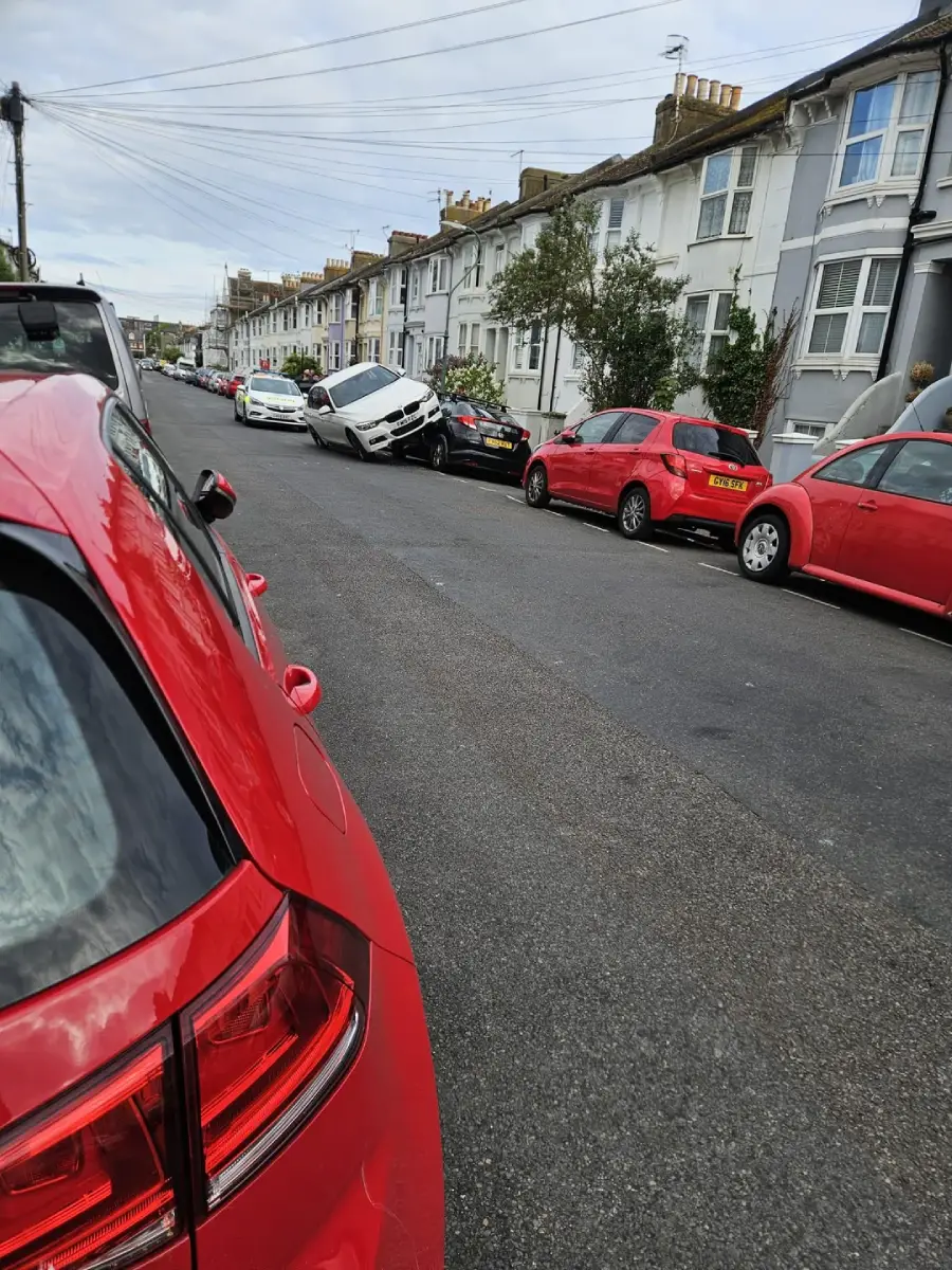 A white car is tilted and partially on top of a black car on the side of a residential street lined with parked cars and terraced houses. The accident appears unusual and dramatic.