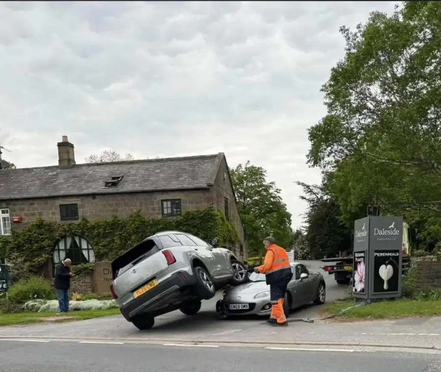 A white SUV is precariously balanced on top of a silver car at the entrance to a rural inn. A person in an orange safety suit inspects the scene, while another person photographs the incident from the sidewalk.