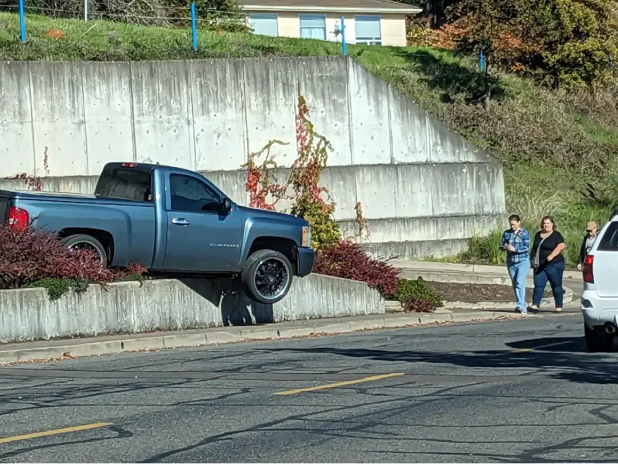 A blue pickup truck is stuck with its front wheels over a concrete curb and the rear wheels still on the road. Three people on the sidewalk look towards the scene, and another vehicle is partly visible on the right.