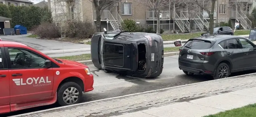 A black SUV is tipped over on its side between a red taxi and a gray SUV on a residential street with apartment buildings in the background.