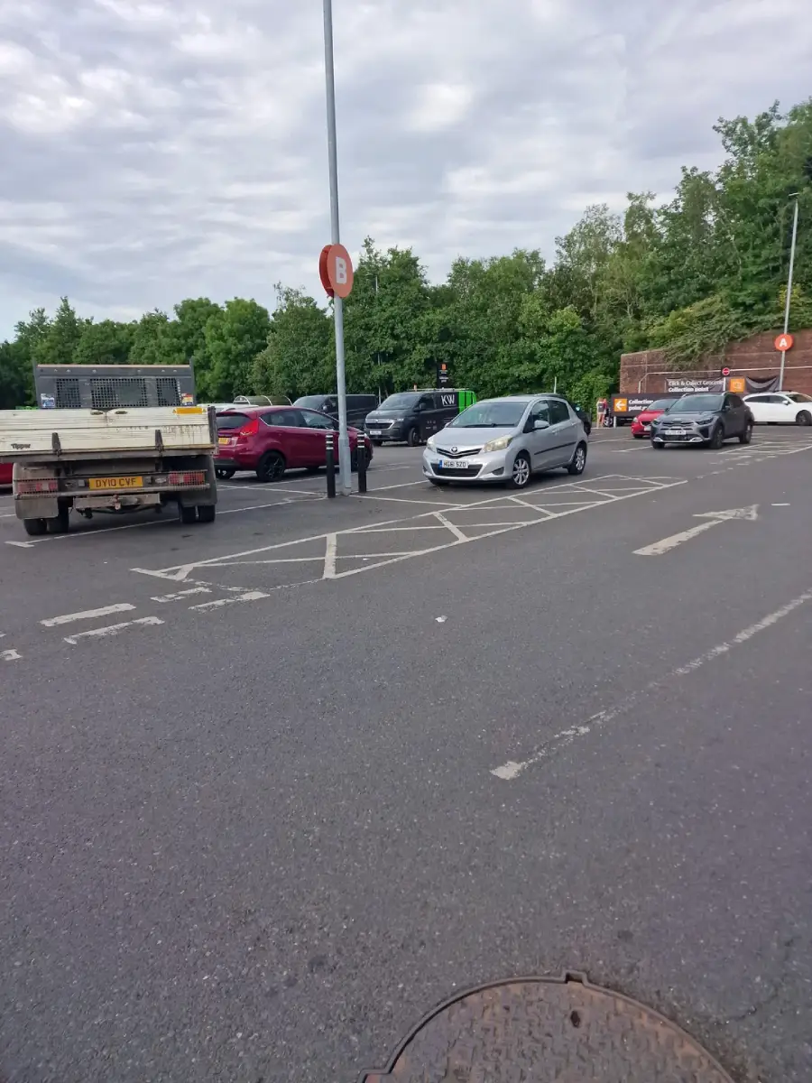 Cars are parked in a parking lot near a "B" section sign, with one silver car parked in a space marked with white diagonal lines. Other vehicles and trees are visible in the background under a cloudy sky.