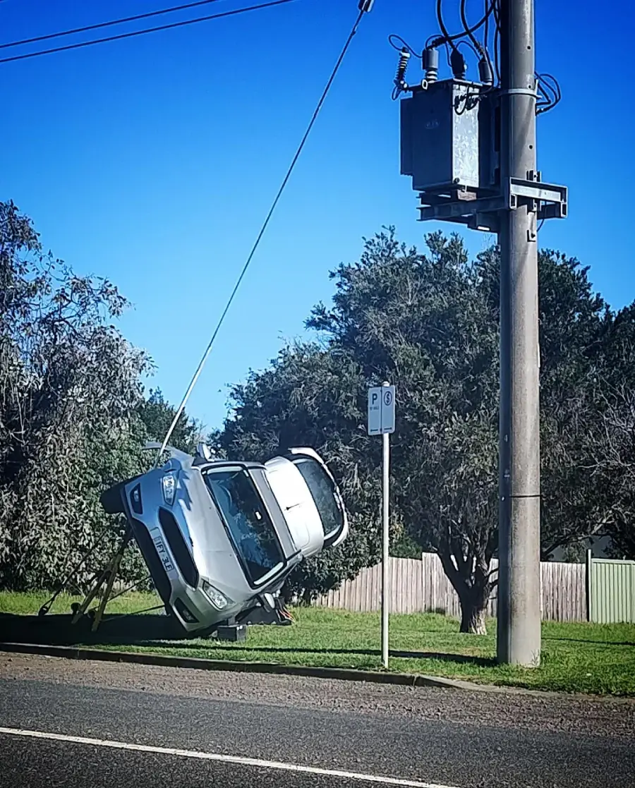 A silver car is hanging sideways, suspended by power lines attached to a utility pole next to a grassy roadside with trees and a wooden fence in the background.