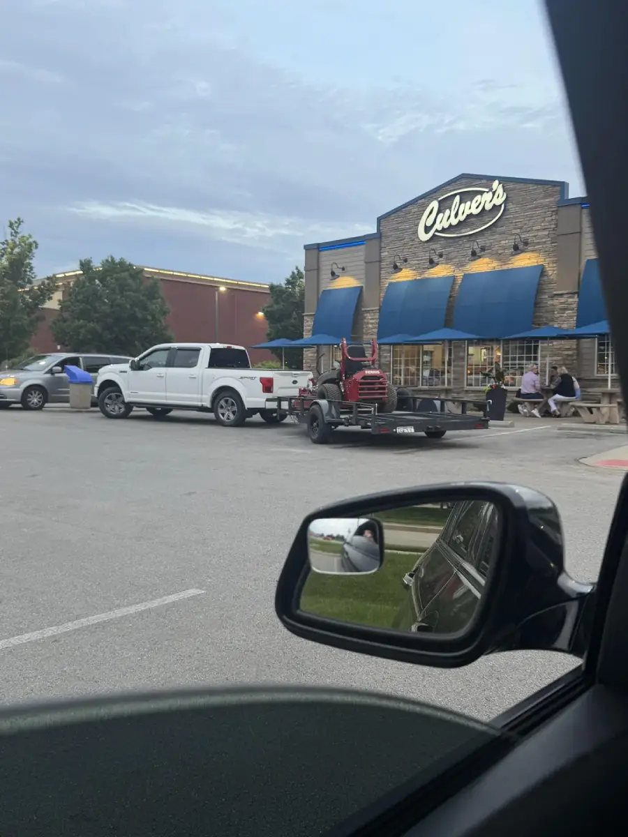 A white pickup truck towing a trailer with a red tractor is parked outside Culver’s restaurant. People are sitting at outdoor tables, and the photo is taken from inside another car, showing a side mirror.
