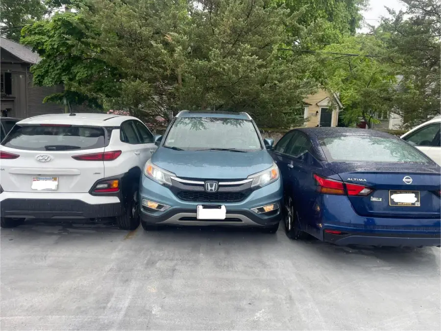 Three cars are parked closely together in a parking lot. The blue car in the center is squeezed tightly between a white car on the left and a dark blue car on the right, leaving very little space between them. Trees and houses are in the background.