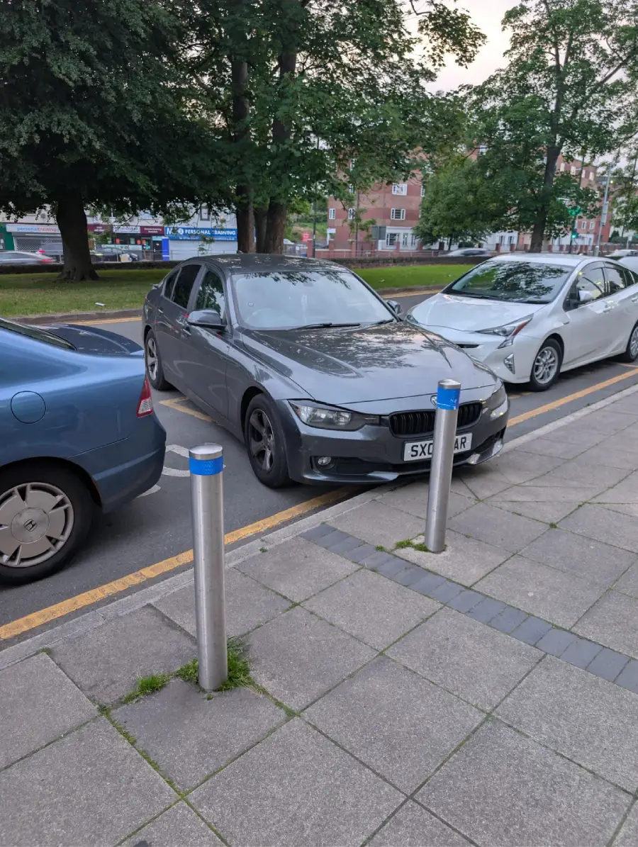 A gray car is parked across two parking spaces, partly on the sidewalk, next to two other cars on a city street lined with trees and apartment buildings.