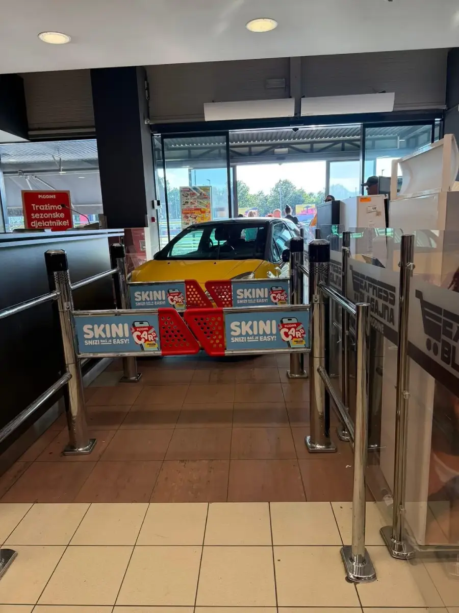A small yellow and red car is parked inside a store, behind metal barriers and near the entrance, blocking the walkway. The scene appears unusual for an indoor retail setting.