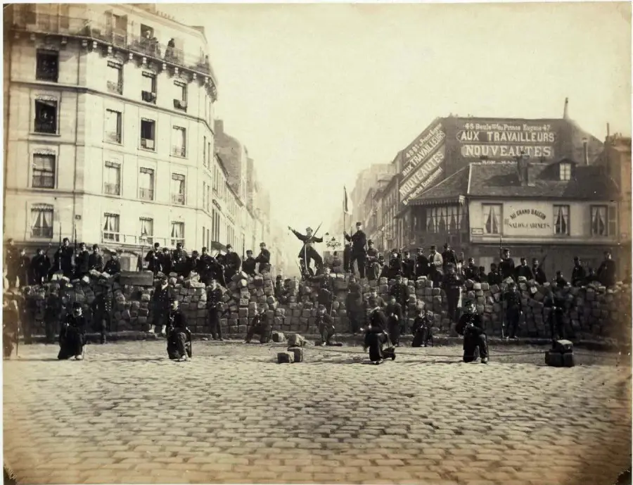 Historic black-and-white photo of people standing and sitting on a large street barricade made of cobblestones and debris, with buildings and shop signs visible in the background.