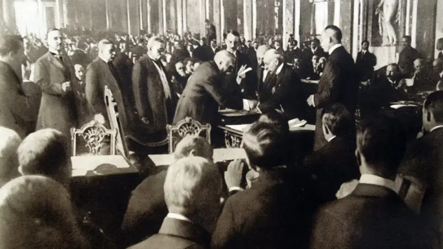 A historic black-and-white photo shows a crowded room of men in formal attire gathered around a table, witnessing officials signing an important document while others observe closely.