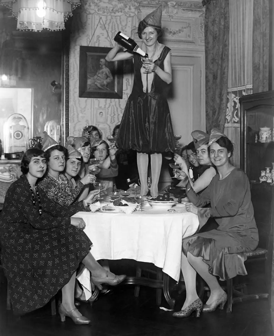 Seven women in party hats sit around a dining table, while one woman stands on the table, pouring wine into her glass. The scene is festive, set in a vintage, ornately decorated room.