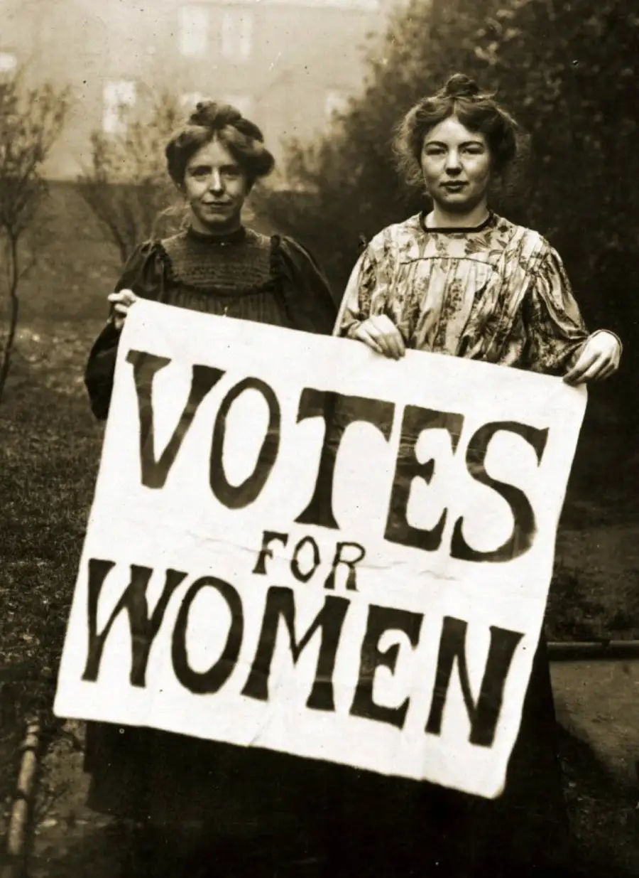 Two women from the early 20th century stand outdoors holding a large handwritten sign that reads “VOTES FOR WOMEN,” supporting the women’s suffrage movement.
