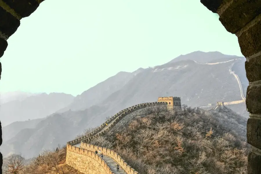 View of the Great Wall of China winding over hills, framed by a stone archway, with mountains in the background and sparse vegetation along the wall.