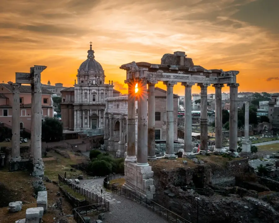Ancient Roman ruins with tall columns and a dome-shaped building in the background, illuminated by a dramatic orange sunset sky, with sunlight shining through the columns.