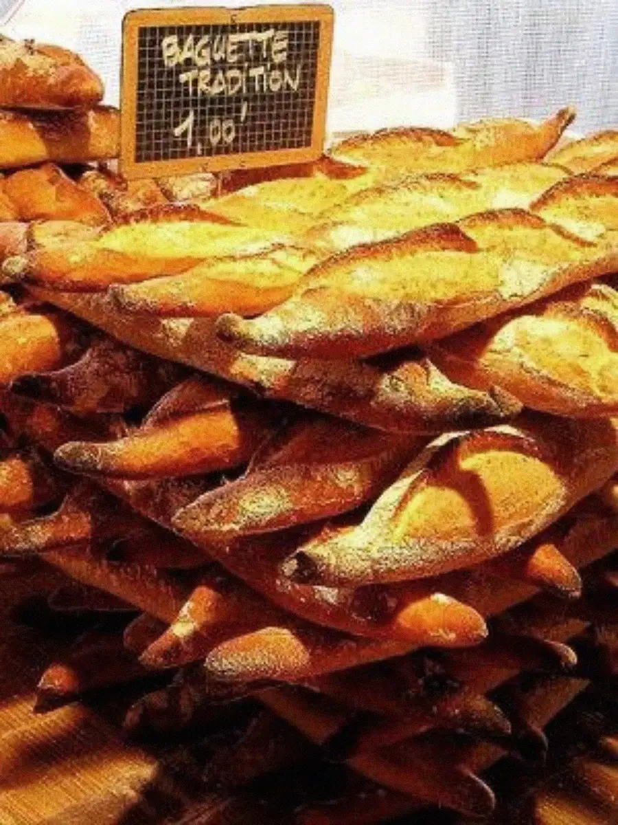 A large stacked display of traditional baguettes is shown at a market. A small chalkboard sign reads "Baguette Tradition 1,00" above the golden-brown, crusty loaves.