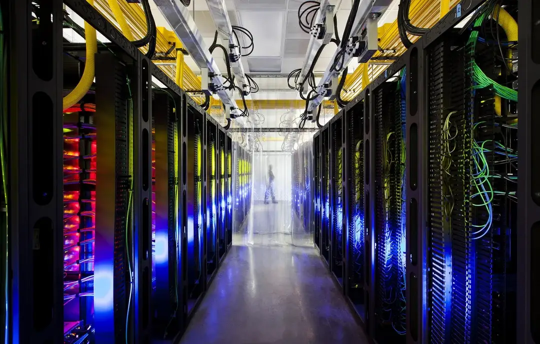 A brightly lit data center corridor lined with server racks on both sides, featuring glowing blue and yellow lights, exposed cables above, and a person silhouetted in the distance through a transparent partition.