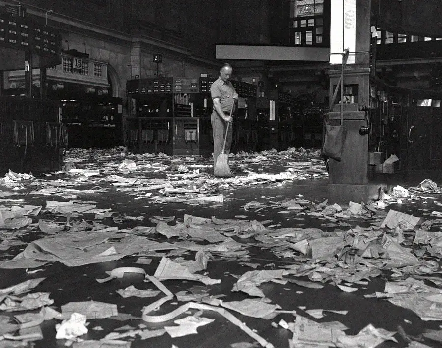 A person sweeps a large floor covered with scattered papers and debris in a dimly lit indoor space with high ceilings and sorting stations.