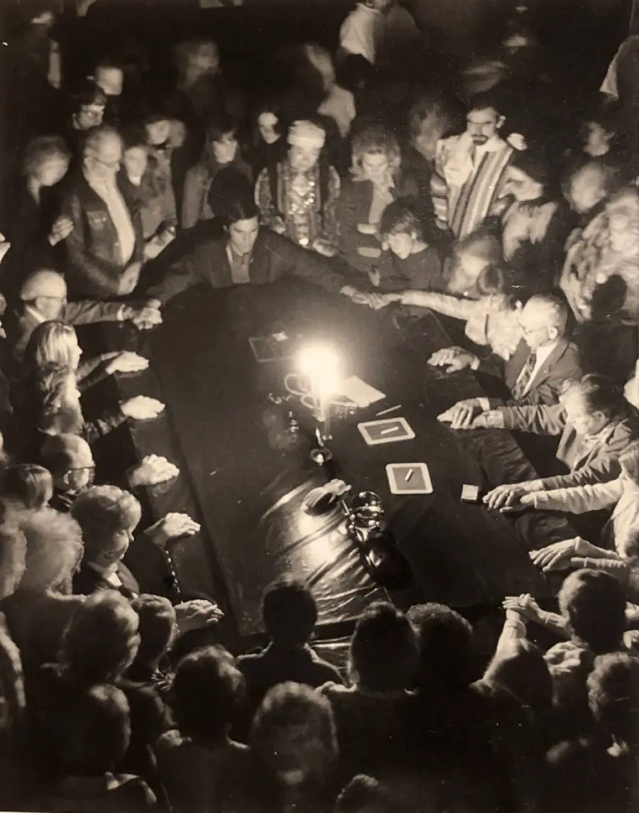 A group of people sit and stand around a dark table, holding hands in a dimly lit room, focused on a single glowing light at the center, creating a mysterious and intense atmosphere.