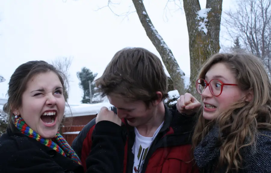 Three teenagers outside in winter, wearing jackets and scarves. One girl on the left is laughing with her mouth wide open, the boy in the middle looks down and covers his face, and the girl on the right looks surprised.