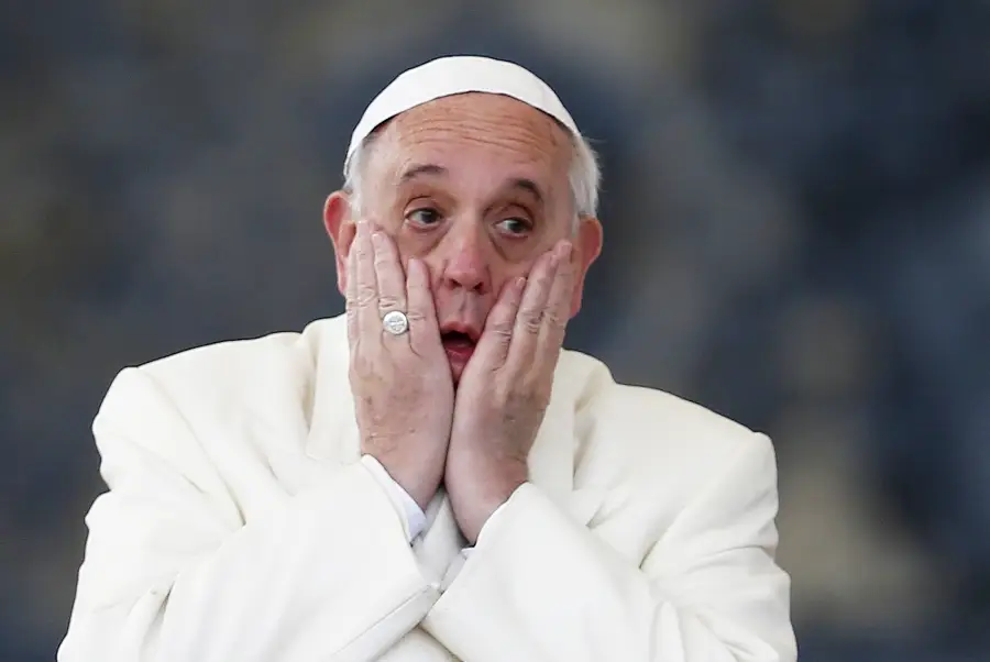 A man wearing a white robe and white cap holds his face with both hands, looking surprised or shocked, against a blurred dark background.