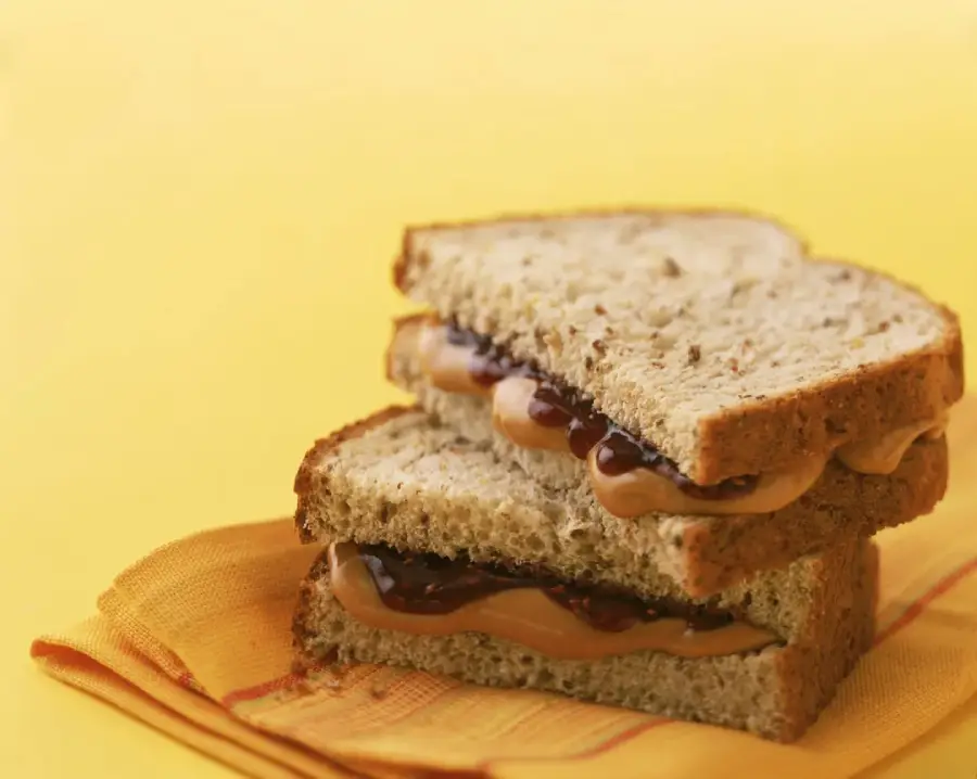 Two halves of a peanut butter and jelly sandwich made with whole grain bread are stacked on top of each other on a folded orange napkin, against a solid yellow background.