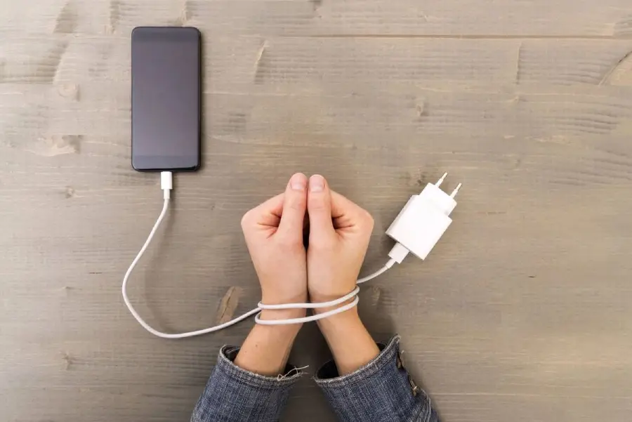 A person's hands are tied together with a phone charger cable, which is also plugged into a smartphone lying on a wooden table, symbolizing dependence on technology.