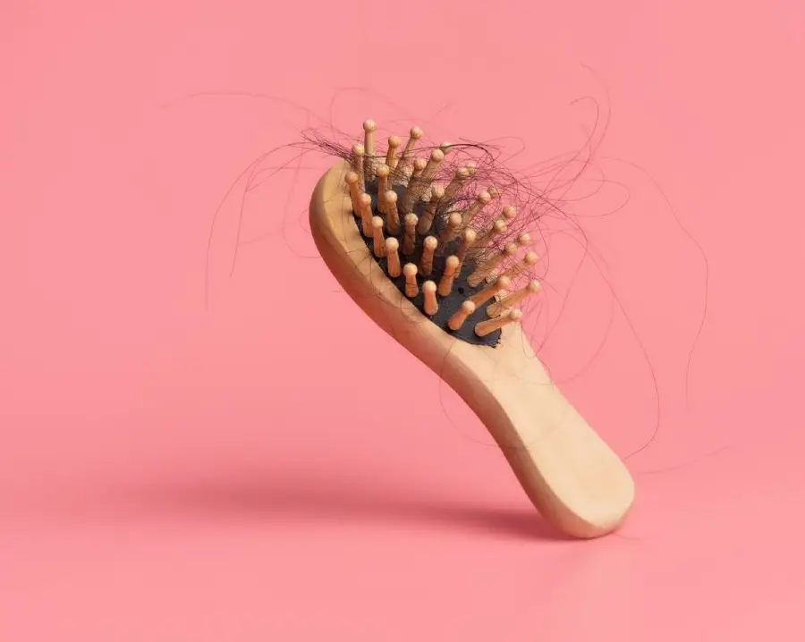 A wooden hairbrush with wooden bristles is shown against a pink background, with a noticeable amount of tangled hair caught in the bristles.