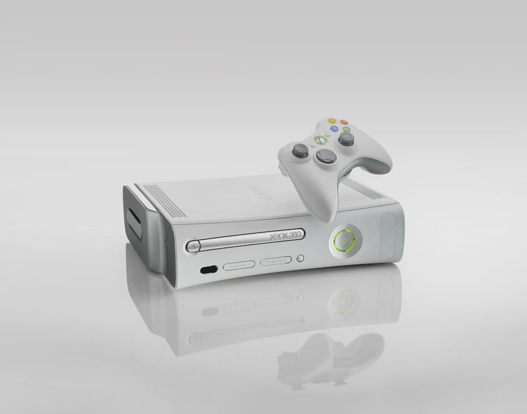 A white Xbox 360 gaming console sits on a reflective surface with a matching wireless controller balanced on top, against a light gray background.