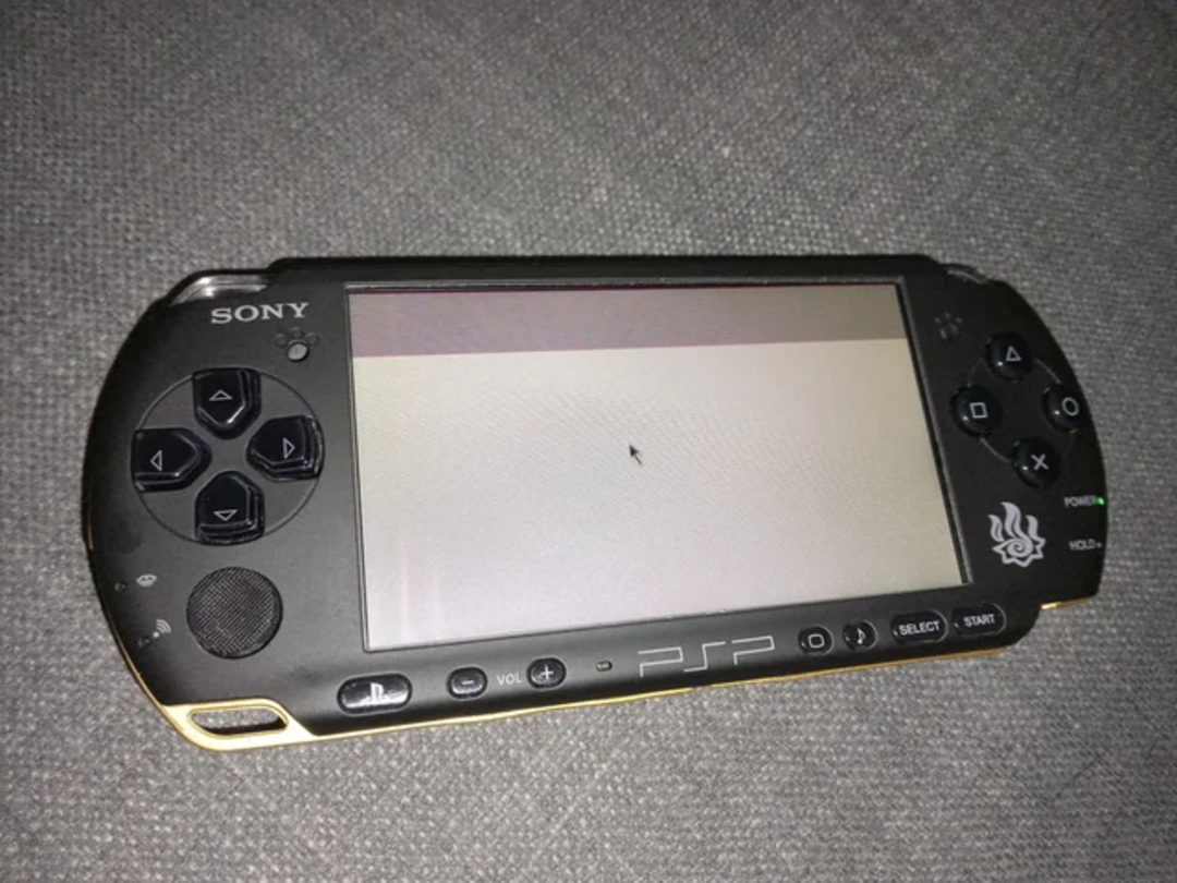 A black Sony PSP handheld gaming console rests on a gray textured surface. Its screen displays a white background with a small black arrow cursor in the center. The buttons and controls are visible.