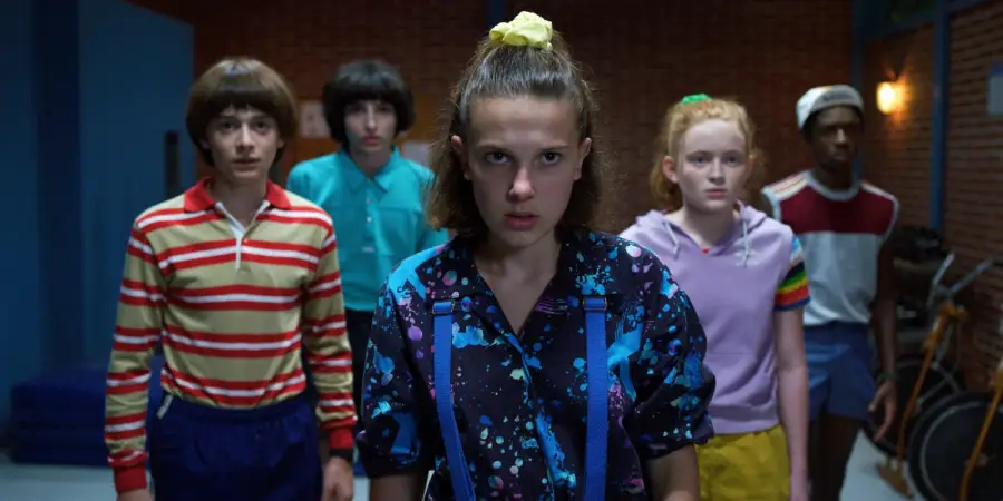 Five teenagers stand close together indoors, looking serious and determined. The girl in front wears a patterned shirt and yellow scrunchie, while the others are positioned behind her, dressed in colorful 1980s-style clothes.