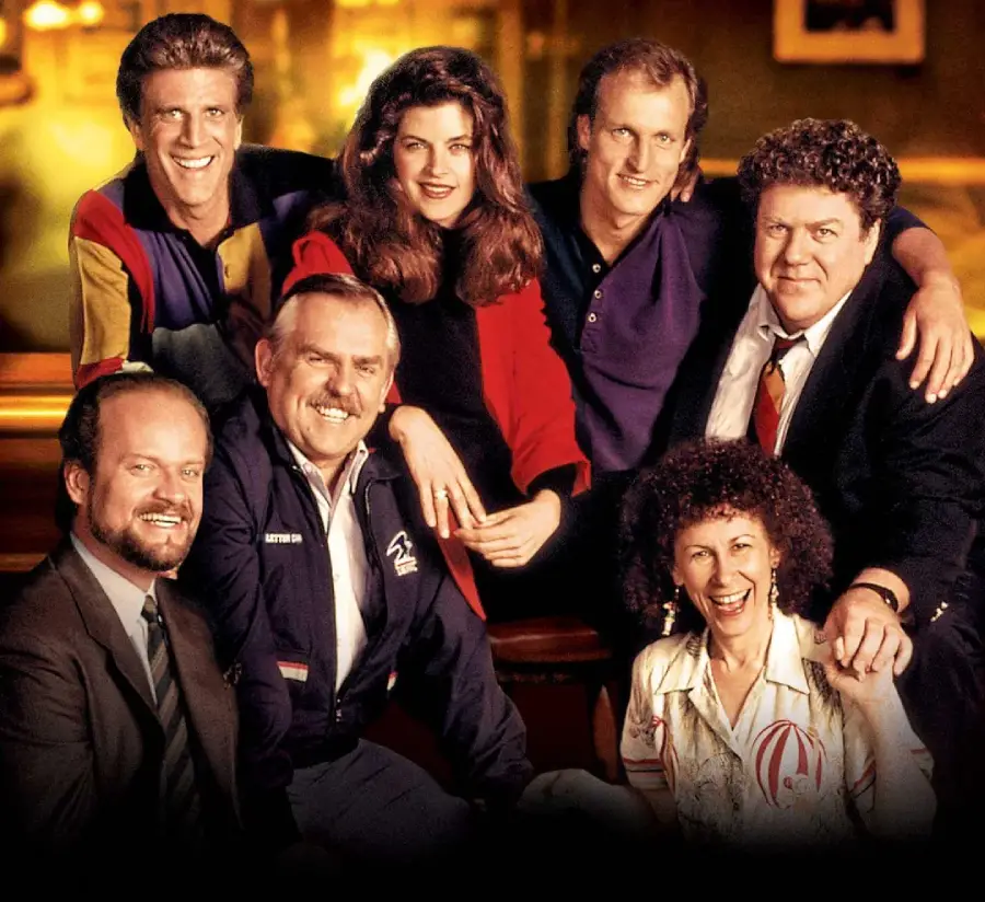 Seven adults, five men and two women, pose together in a warmly lit bar setting, smiling at the camera. They are dressed in casual and semi-formal 1980s-style clothing.