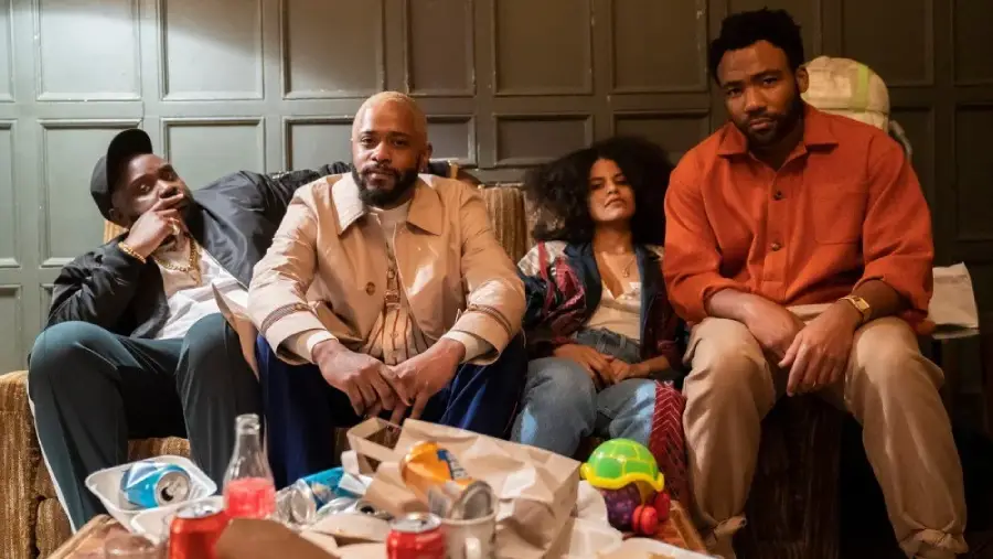 Four people sit on a couch in a dimly lit room, surrounded by clutter and empty food containers. They appear relaxed but thoughtful, with neutral or slightly serious expressions.