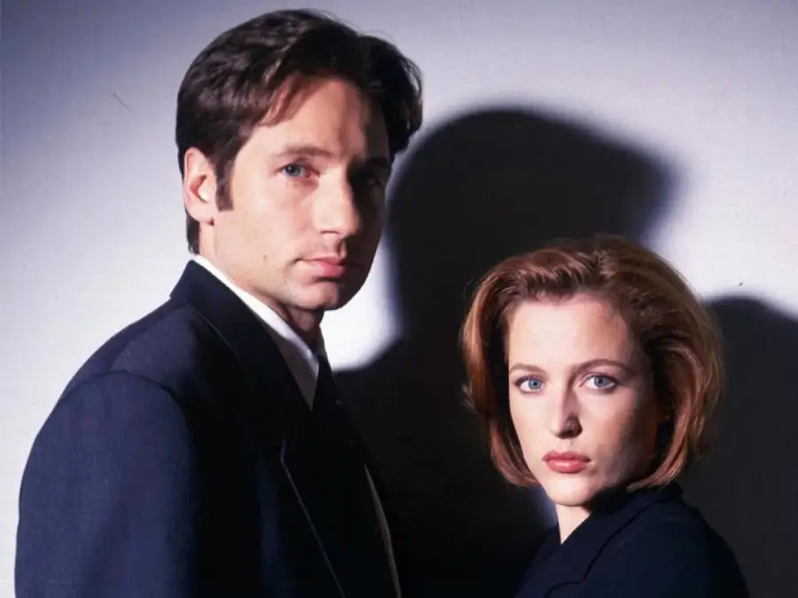 A man and woman in dark suits stand side by side against a light background, both looking seriously at the camera. The lighting casts a strong shadow on the wall behind them.