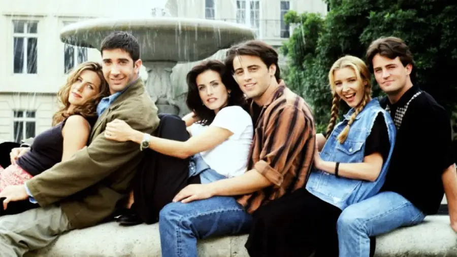 Six young adults sit closely together on the edge of a fountain outdoors, smiling and leaning on each other, with a large building and trees in the background.