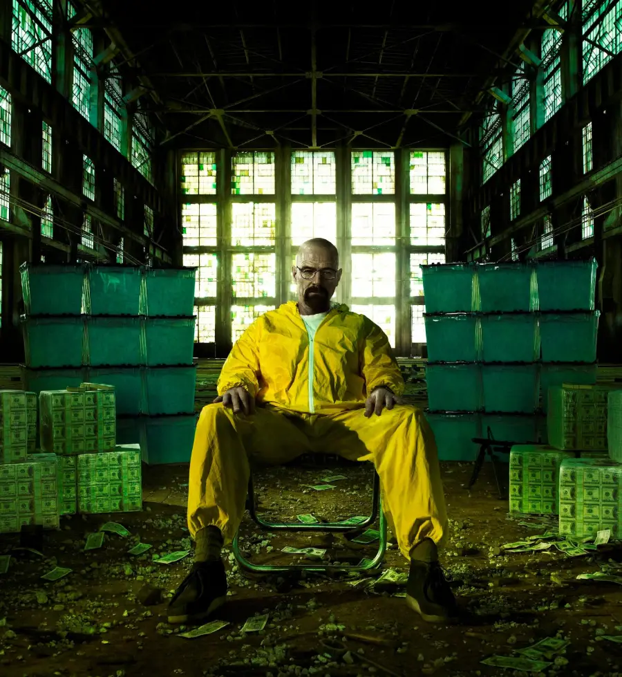 A man in a yellow hazmat suit sits in a chair inside a large, abandoned warehouse, surrounded by stacks of blue containers and piles of cash scattered on the floor. Green light filters through tall windows behind him.