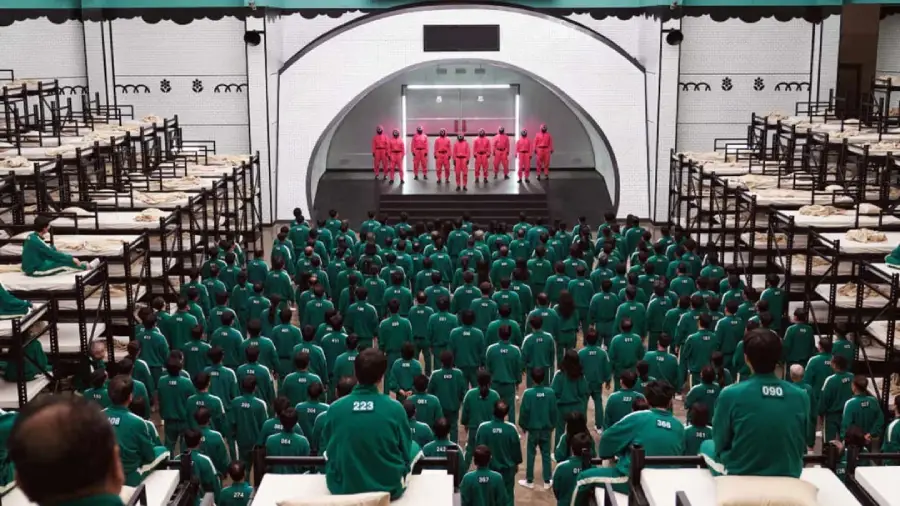 Hundreds of people in green tracksuits stand in rows facing a stage, where masked guards in pink uniforms stand in a line; the room is filled with stacked bunk beds.
