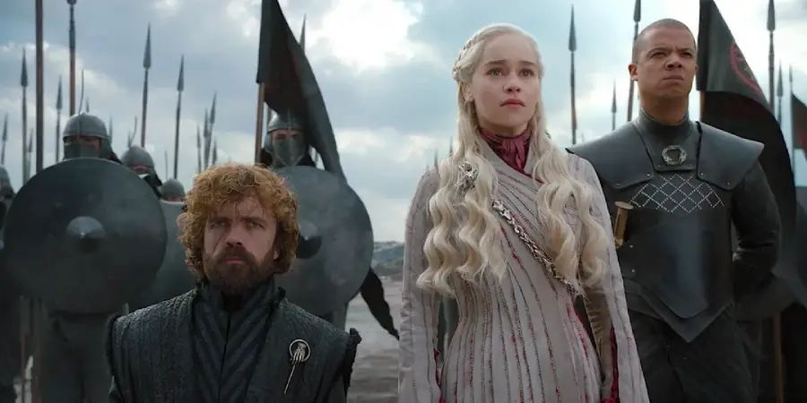 Three characters stand side by side in front of an army with spears and shields. The woman in the center has long blonde hair and wears a light-colored outfit. The two men beside her wear dark clothing and serious expressions.